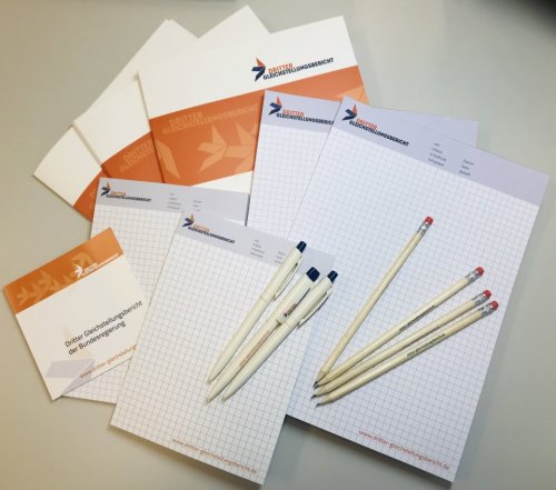 Foto von Blöcken, Bleistiften und Broschüren der Geschäftsstelle Dritter Gleichstellungsbericht mit dem design der Geschäfsstelle in orange und dunkelblau.