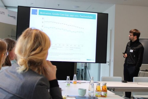 Foto von Dr. Kai-Uwe Müller beim Vortrag mit Zuhörenden an Konferenztisch. In der Mitte des Bildes die Leinwand mit Vortragsfolie zum Thema Gender-Care-Gap.