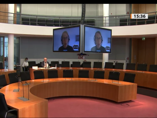 Foto eines Konferenzraums im Bundestag. links im Hintergrund sitzt eine Person. In der Mitte des Raums hängen große Bildschirme über die ein Videoanruf übertragen wird.
