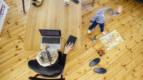 Foto aus Vogelperspektive von Person, die mit Laptop an einem Tisch sitzt, danaben spielt ein Kind auf dem Boden